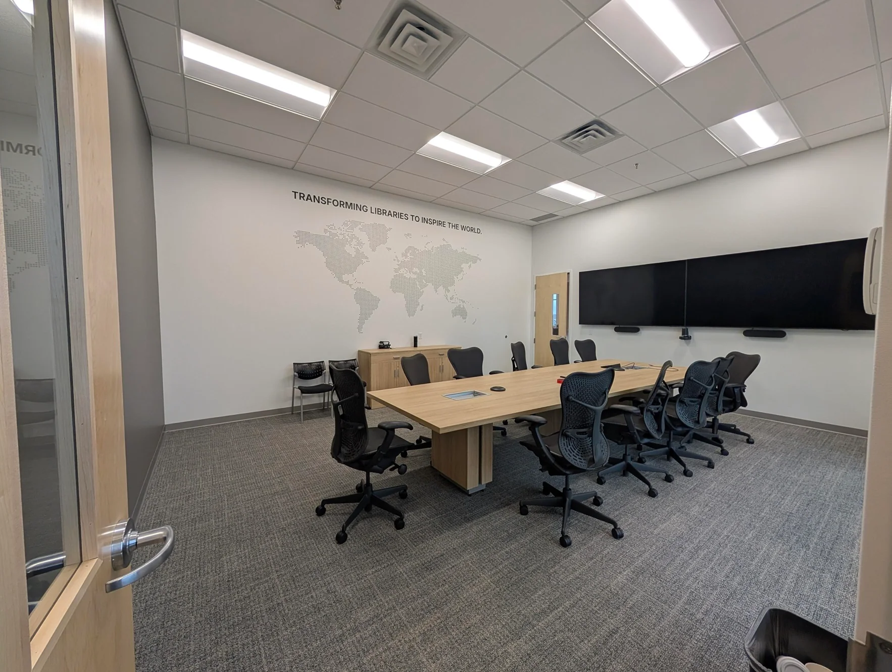 A modern meeting room with a large wooden conference table, ergonomic chairs, and dual wall-mounted screens, featuring a world map decal on the wall.
